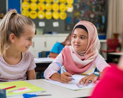 Young arab girl with hijab doing exercise with her bestfriend at international school. Asian muslim school girl sitting near her classmate during lesson. Multiethnic elementary students in classroom.